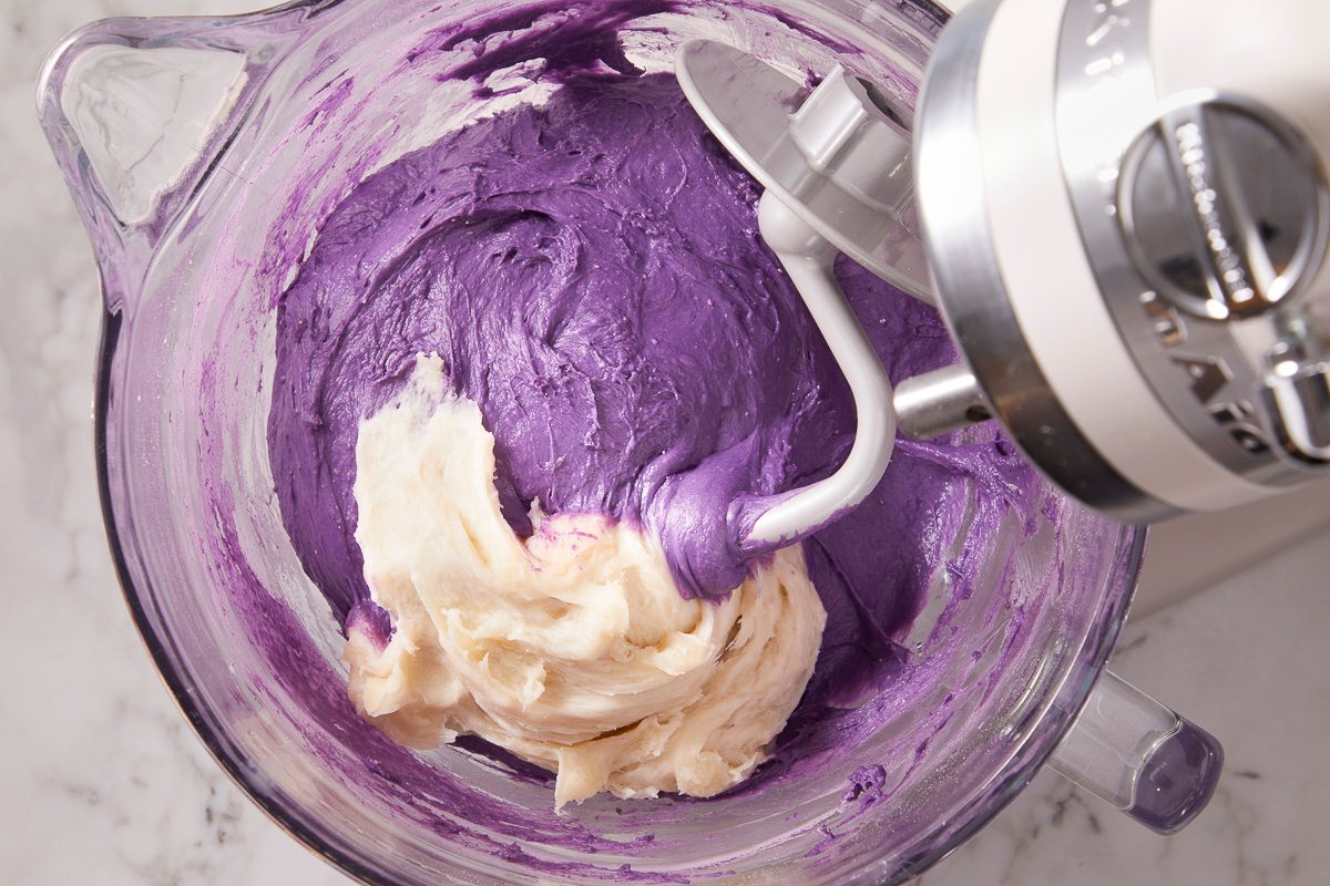 Overhead horizontal image of dough being mixed in a clear glass stand mixer bowl. A light-colored dough is being incorporated into a vibrant purple ube dough by the mixer's paddle attachment.