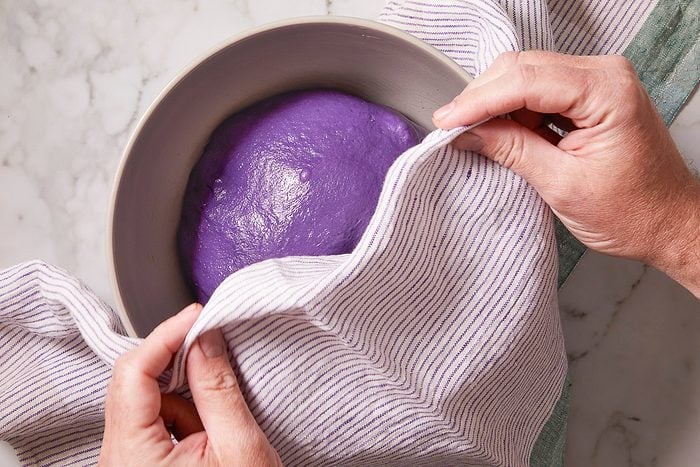 Overhead horizontal image of hands uncovering a bowl of vibrant purple ube dough with a striped kitchen towel. The dough is shown after rising, ready for the next step in the baking process.