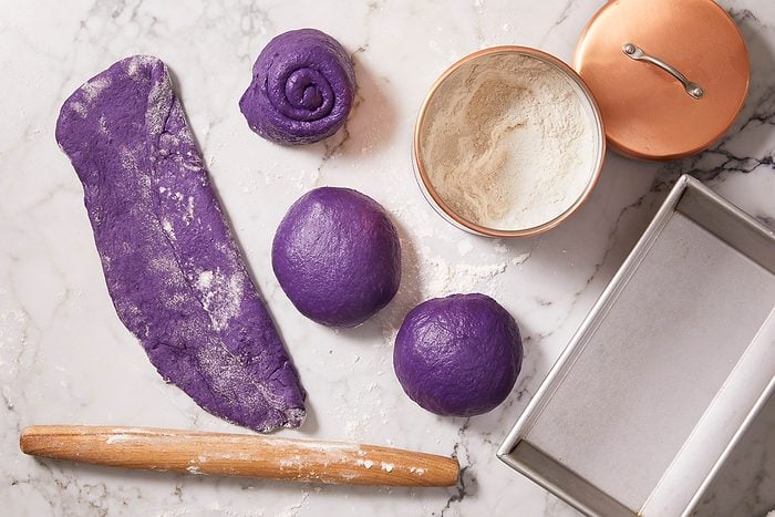 Overhead horizontal image of vibrant purple ube bread dough being prepared on a marble surface dusted with flour. Various portions of the dough are shown, including one rolled flat, several shaped into balls, and one swirled, alongside a wooden rolling pin, a metal tin filled with flour, and an empty loaf pan.