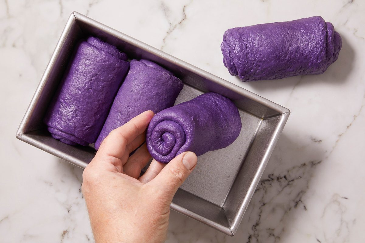 Overhead horizontal image of a hand placing a rolled-up piece of vibrant purple ube bread dough into a metal loaf pan. Two other rolled dough pieces are already arranged in the pan, with an additional piece resting beside it on a marble surface.
