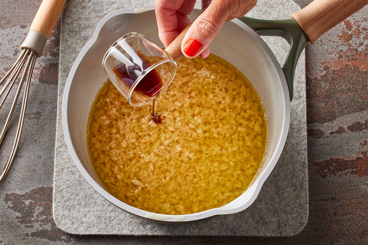 Overhead horizontal image of a hand pouring a dark liquid from a small measuring spoon into a light gray pot. The pot contains finely minced garlic simmering in golden oil, with a metal whisk resting on a gray mat nearby. | [Original] Melt butter in a saucepan or wok over medium heat. Add garlic and cook for 2 minutes, being careful not to brown. Add fish sauce, oyster sauce, sugar and Maggi seasoning. Stir to combine, then remove from heat.