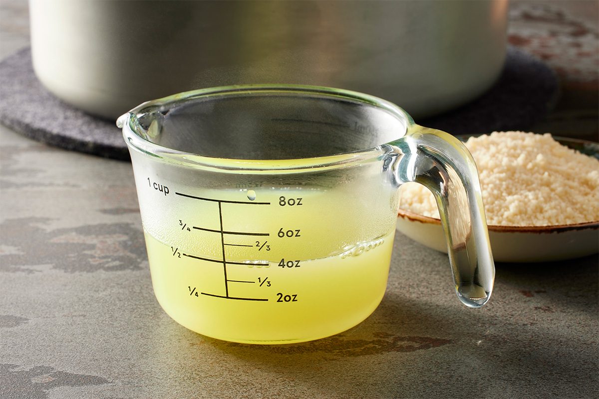 Eye Level horizontal image of a glass measuring cup holding approximately 4oz of a pale yellow, steaming liquid. In the background, a large pot is partially visible, and a bowl filled with grated cheese rests to the right. | [Original] Meanwhile, bring a pot of water to boil. Cook fresh chow mein noodles according to package directions. Drain; reserving 1/2 cup cooking water.