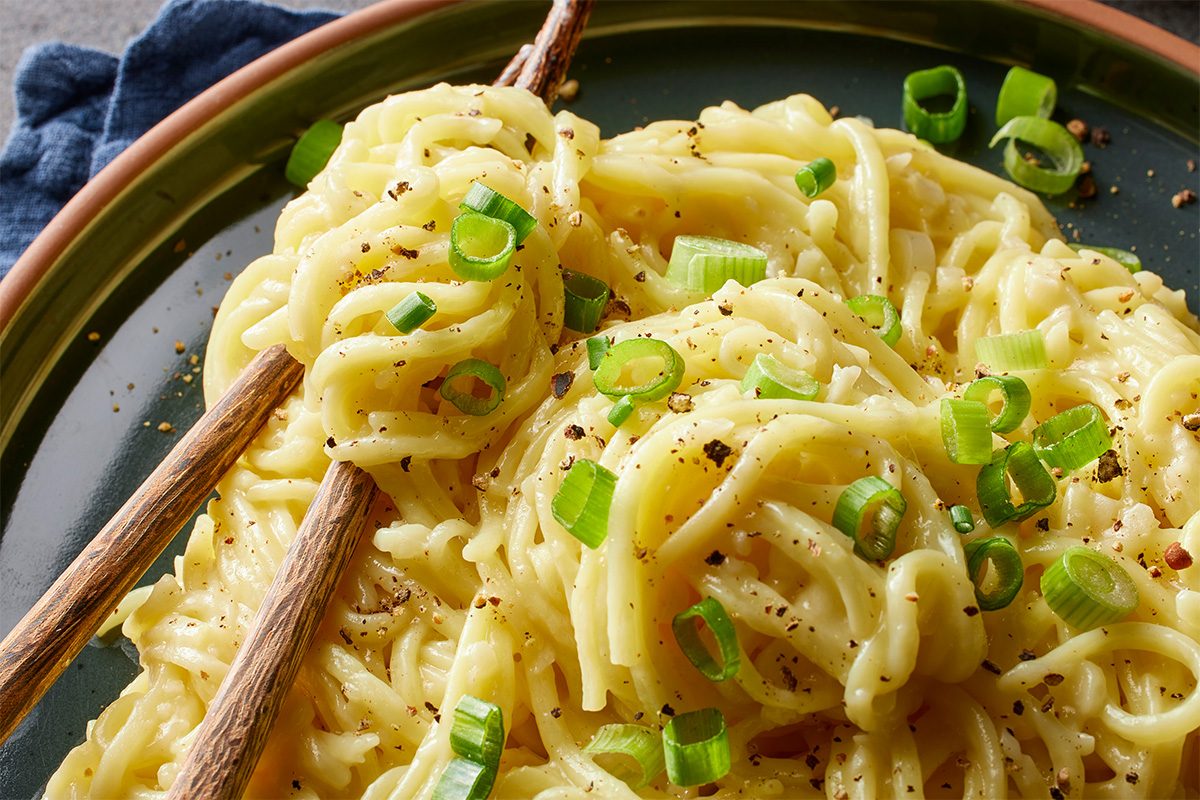 Eye Level horizontal image of a close-up of creamy noodles, generously garnished with chopped green onions and freshly cracked black pepper. A pair of chopsticks rests on the inviting dish, served on a green plate.