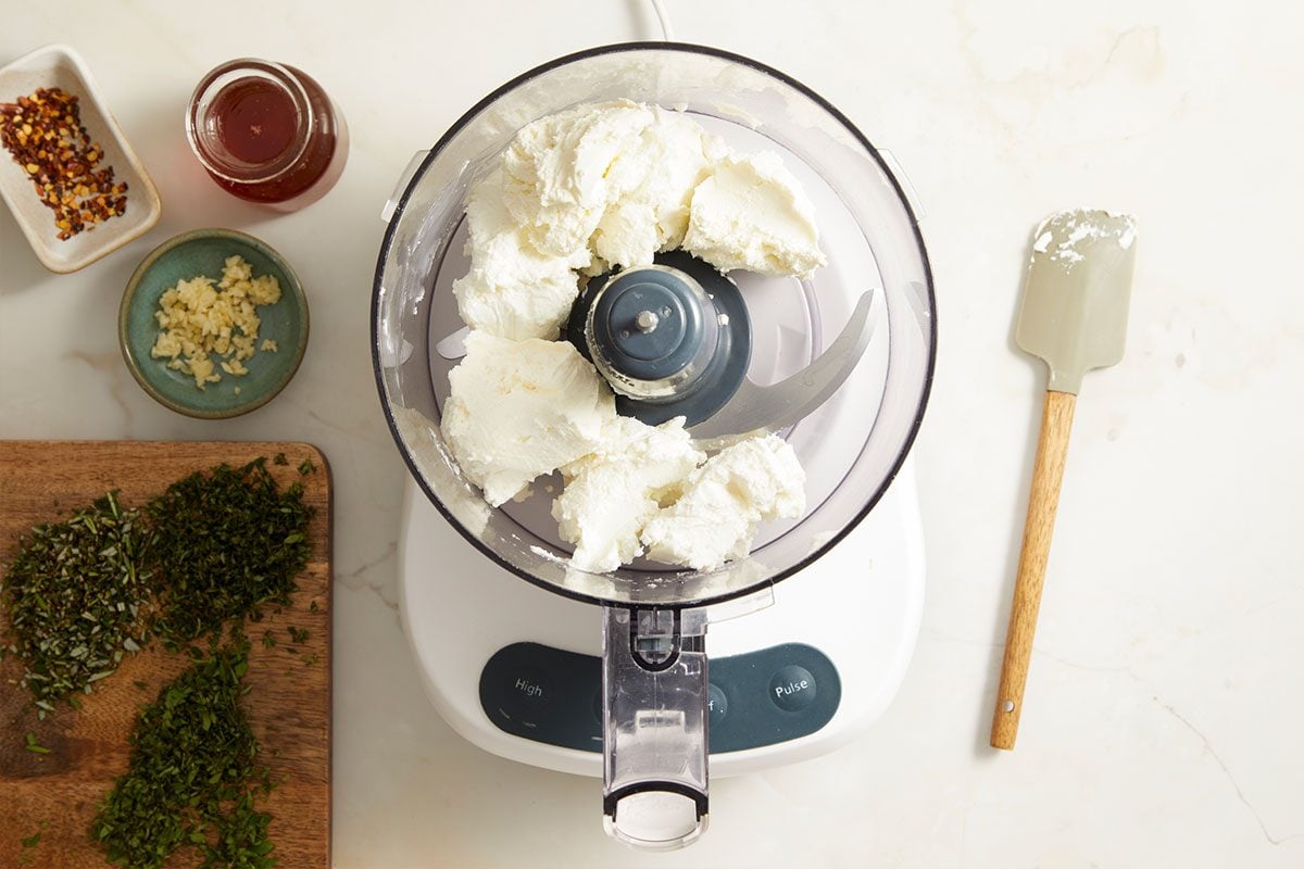 A food processor on a kitchen counter with an assortment of ingredients arranged around it for cooking. 