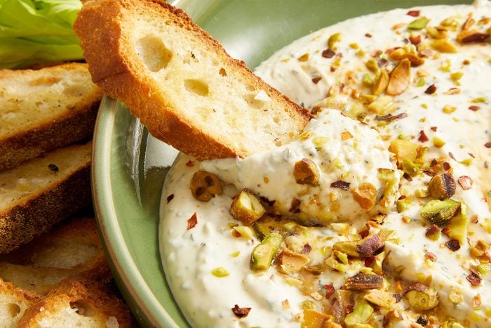 A bowl of creamy dip garnished with crunchy nuts and bread, served with extra bread on the side.