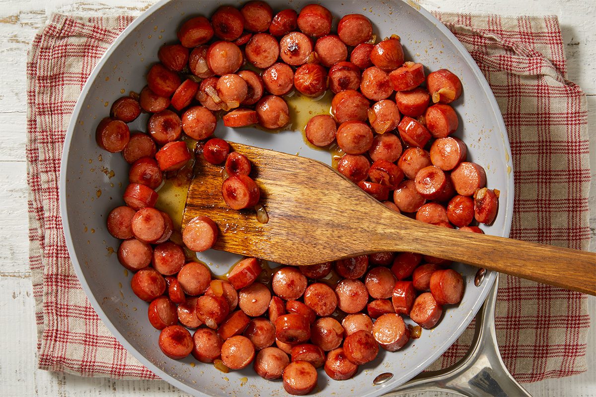 Overhead horizontal image of sliced hot dogs and diced onions cooking in a light gray frying pan. A wooden spatula rests among the sizzling ingredients, which are coated in a rich, glistening sauce. The pan is placed on a red and white plaid dish towel on a light wooden background.