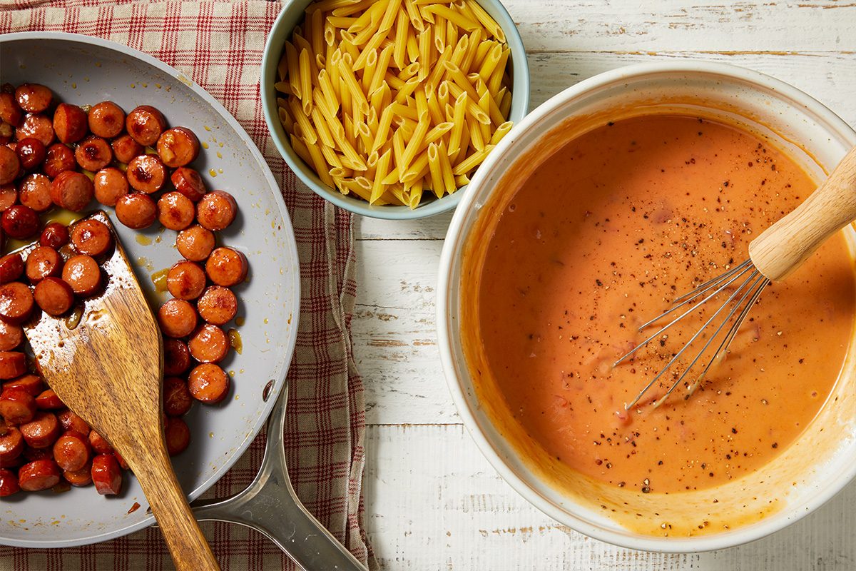 Overhead horizontal image of ingredients arranged on a white wooden surface with a red and white checkered cloth. Sliced cooked sausage is in a pan with a wooden spatula, alongside a bowl of uncooked penne pasta and a bowl of creamy orange sauce with black pepper and a whisk. These components are prepared for a recipe.
