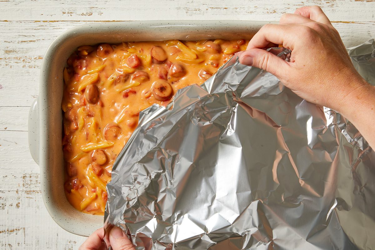 Overhead horizontal image of hands covering a casserole dish filled with a creamy pasta and hot dog mixture with aluminum foil. The dish contains elbow macaroni, sliced hot dogs, and a cheese-like sauce, ready for baking.
