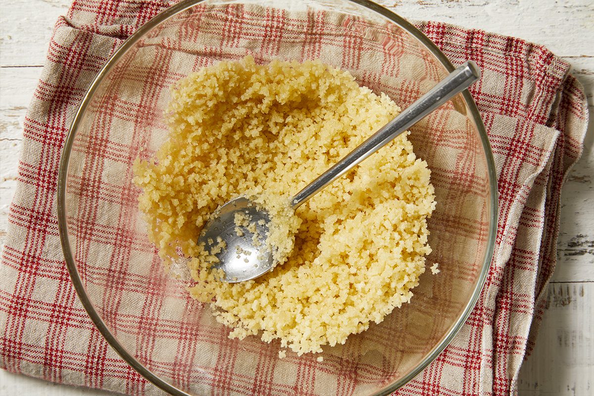 Overhead horizontal image of fluffy, cooked couscous in a clear glass bowl. A silver spoon rests in the couscous, ready for serving or mixing. The bowl is placed on a white wooden surface, with a red and white plaid kitchen towel underneath.