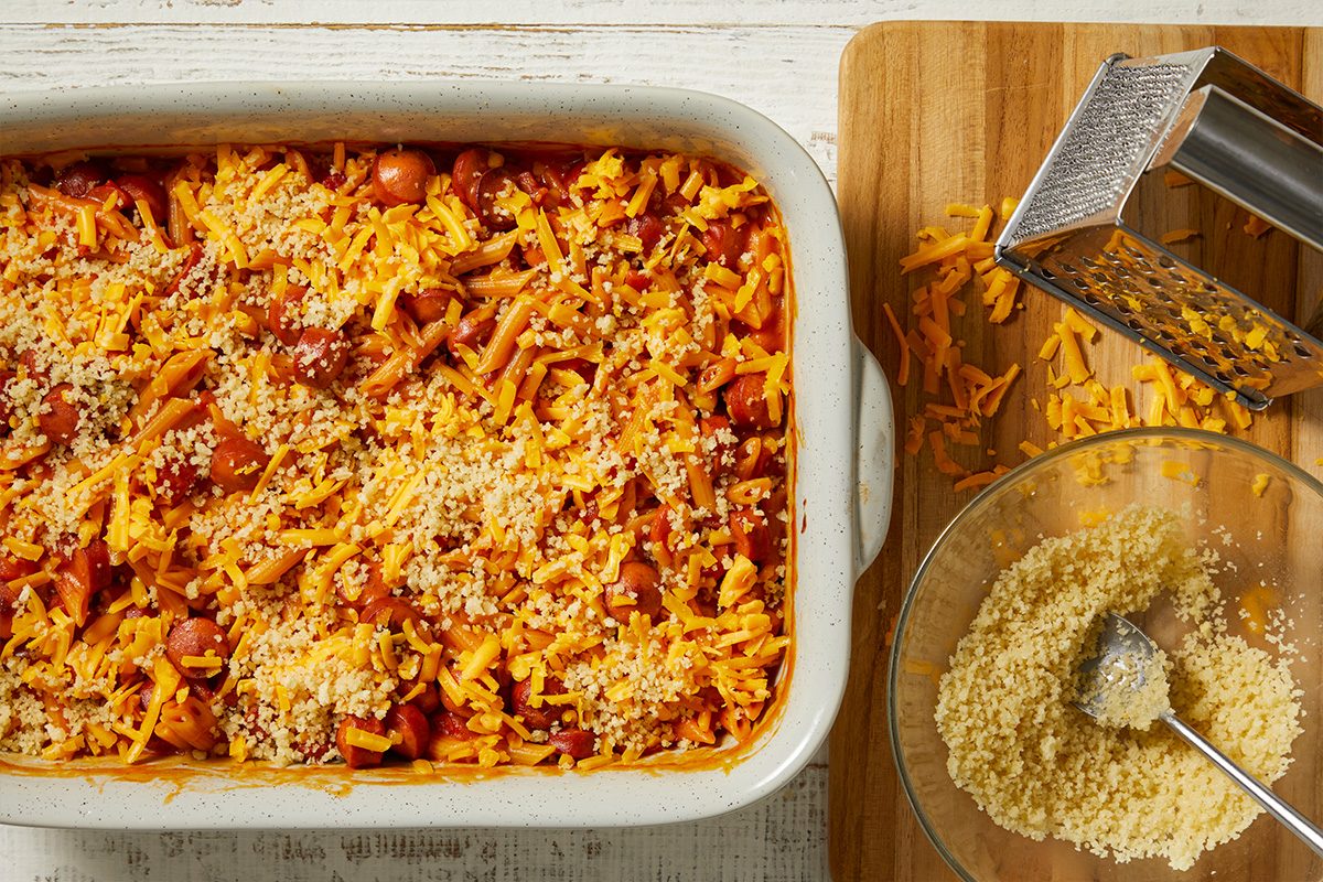 Overhead horizontal image of a casserole in a white baking dish. The dish contains pasta and sliced hot dogs, generously topped with shredded cheddar cheese and breadcrumbs. To the right, a wooden cutting board holds a box grater and some loose shredded cheese, alongside a glass bowl filled with breadcrumbs and a spoon.