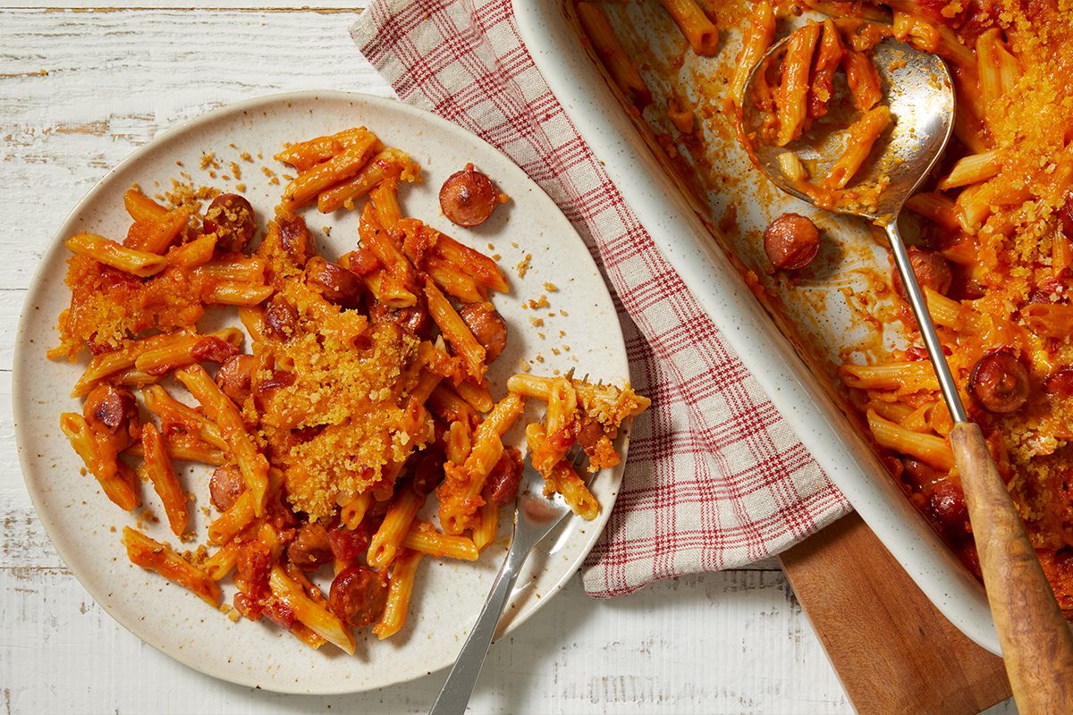 Overhead horizontal image of a serving of penne pasta with sliced hot dogs and a golden-brown breadcrumb topping on a speckled plate. A fork rests in the pasta on the plate, next to a white baking dish containing more of the saucy pasta bake with a serving spoon.