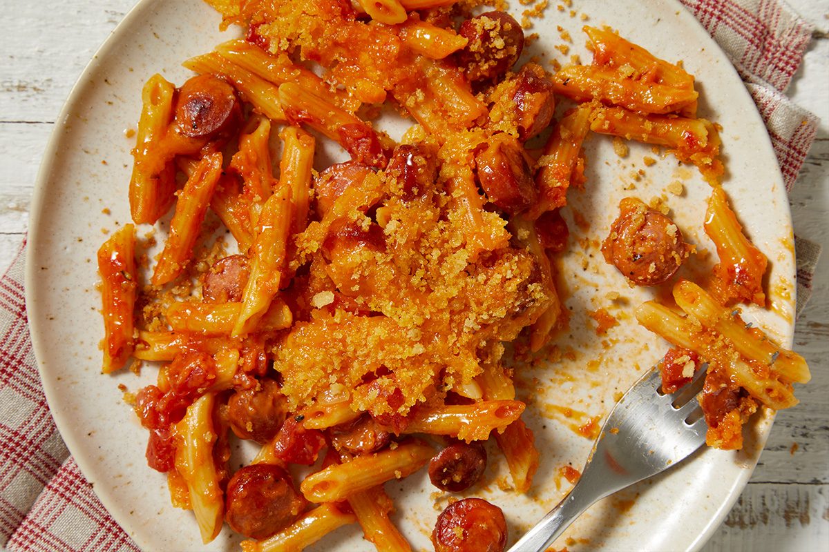 Overhead horizontal image of a hearty baked pasta dish on a speckled cream plate, featuring penne pasta, sliced sausage, a rich tomato sauce, and a golden, crunchy breadcrumb topping. A silver fork rests on the right side of the plate, which is set on a distressed white wooden surface with a checkered napkin.