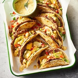 A tray of crispy tacos filled with ground beef, chopped lettuce, diced tomatoes, pineapple, and a creamy yellow sauce, with a small bowl of sauce and a spoon on the side.