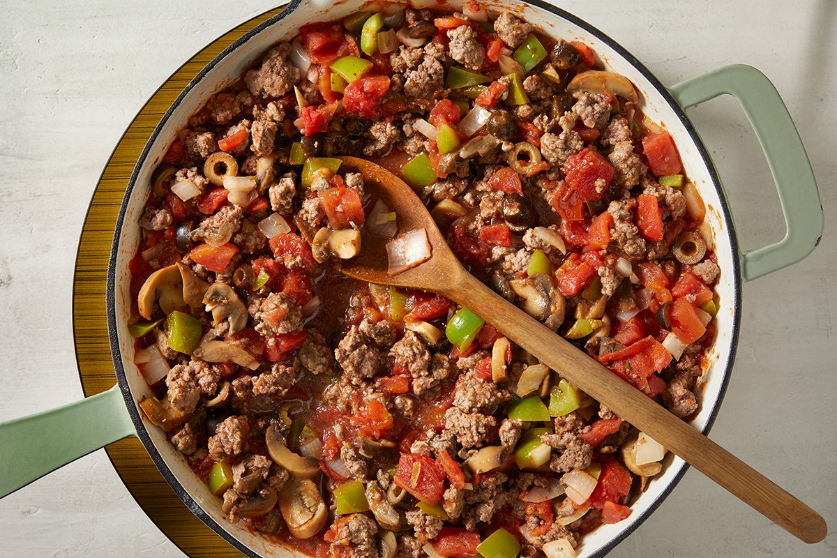 A large skillet filled with a colorful mixture of ground meat, diced tomatoes, green bell peppers, onions, and sliced olives. A wooden spoon rests in the pan, ready for serving.