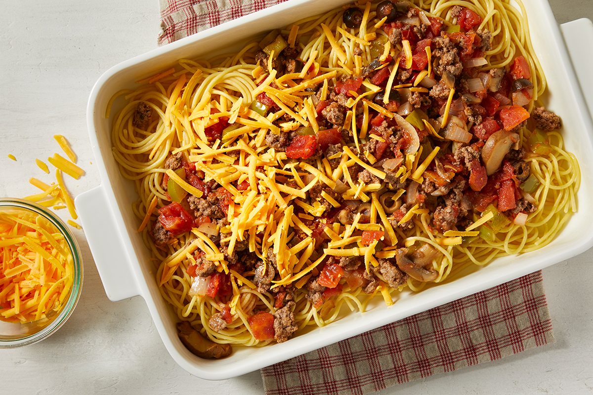 A white baking dish filled with cooked spaghetti, ground beef, diced tomatoes, mushrooms, and shredded cheddar cheese on top, placed on a red and white checkered cloth. A small bowl of cheddar cheese is beside the dish.