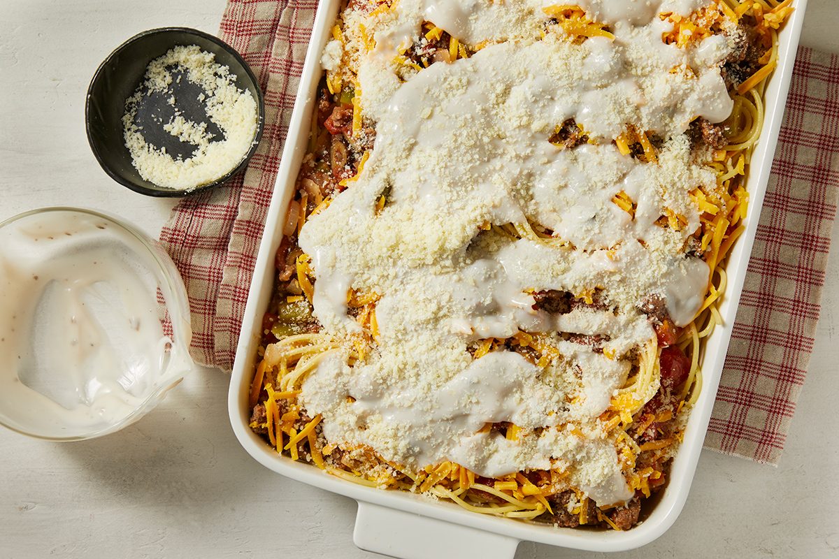 A casserole dish filled with baked spaghetti topped with melted cheese and creamy white sauce sits on a red-checked cloth, with a bowl and small dish of grated cheese nearby.