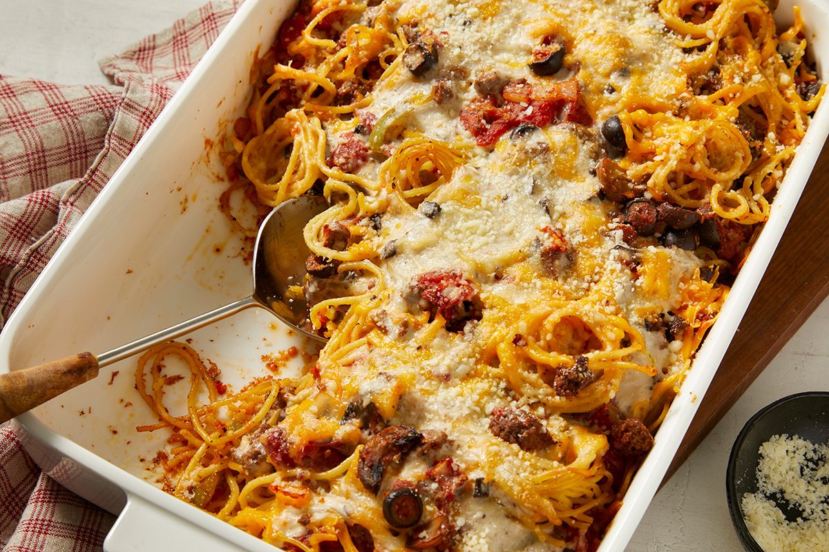 A baked spaghetti casserole topped with melted cheese, tomato sauce, and olives sits in a white dish with a serving spoon. A red-and-white cloth and a small bowl of grated cheese are nearby.