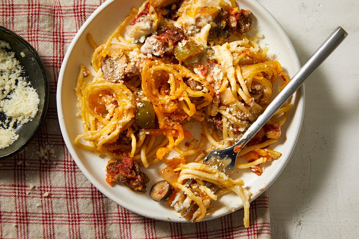A plate of spaghetti with tomato sauce, vegetables, and grated cheese sits on a red and white checkered cloth. A fork rests on the plate, and a bowl of grated cheese is nearby.