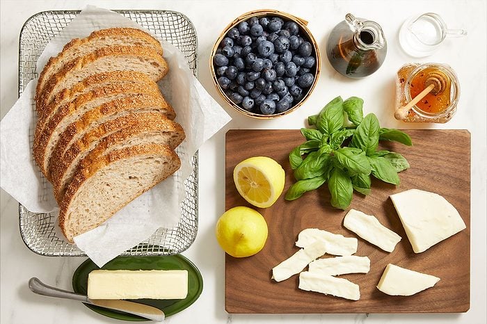 Overhead shot of sliced rustic bread arranged on a cooling rack, with fresh blueberries, basil leaves, lemons, sliced mozzarella cheese, butter, honey, and balsamic vinegar prepared on a wooden cutting board and light surface for recipe preparation.