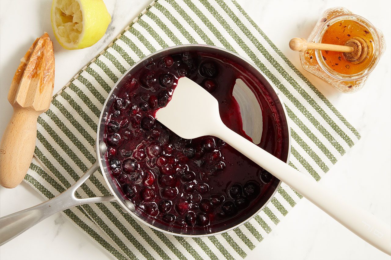 Overhead shot of a saucepan filled with simmering blueberries cooking into a fruit compote, stirred with a white silicone spatula. A lemon juicer, honey jar with a honey dipper, and a striped kitchen towel are arranged nearby on a light surface.