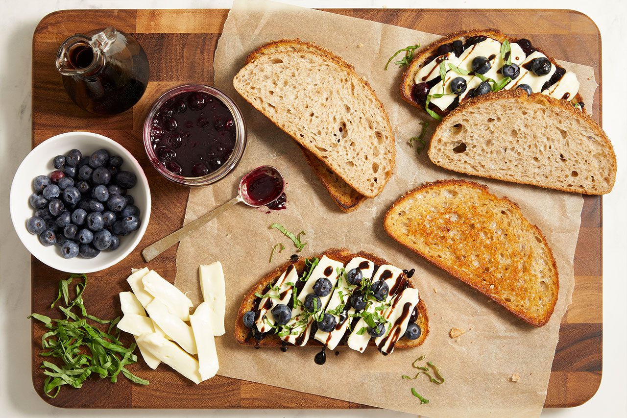 Overhead shot of toasted crostini prepared with creamy cheese, blueberry compote, fresh blueberries, and basil, arranged on a wooden serving board alongside sliced rustic bread, fresh mozzarella, blueberries, blueberry compote, and balsamic vinegar.