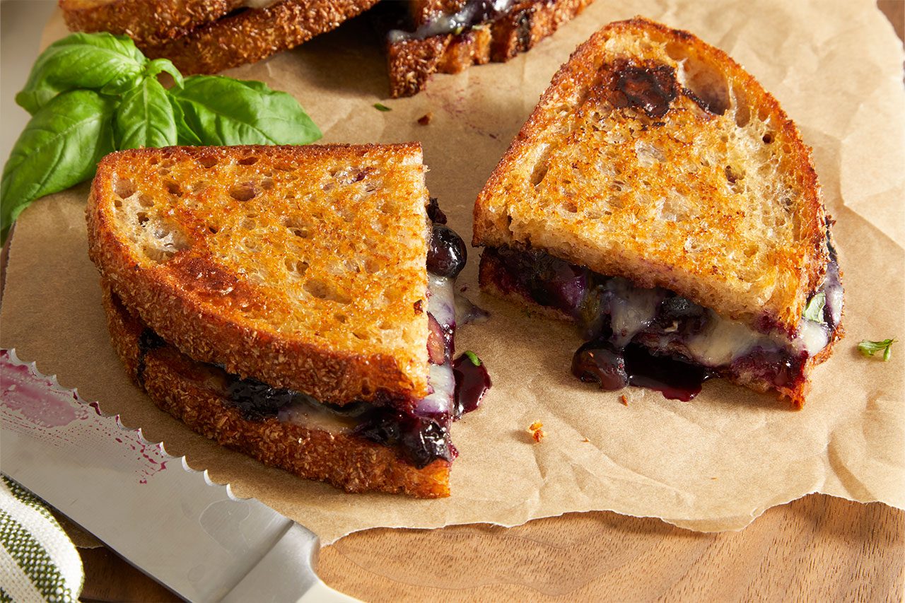 3/4 angle view shot of Balsamic Blueberry Brie Grilled Cheese sandwiches with golden, crispy toasted bread filled with melted Brie cheese and warm blueberry compote, arranged on parchment paper with fresh basil and a serrated knife nearby, highlighting the gooey filling and crisp texture.