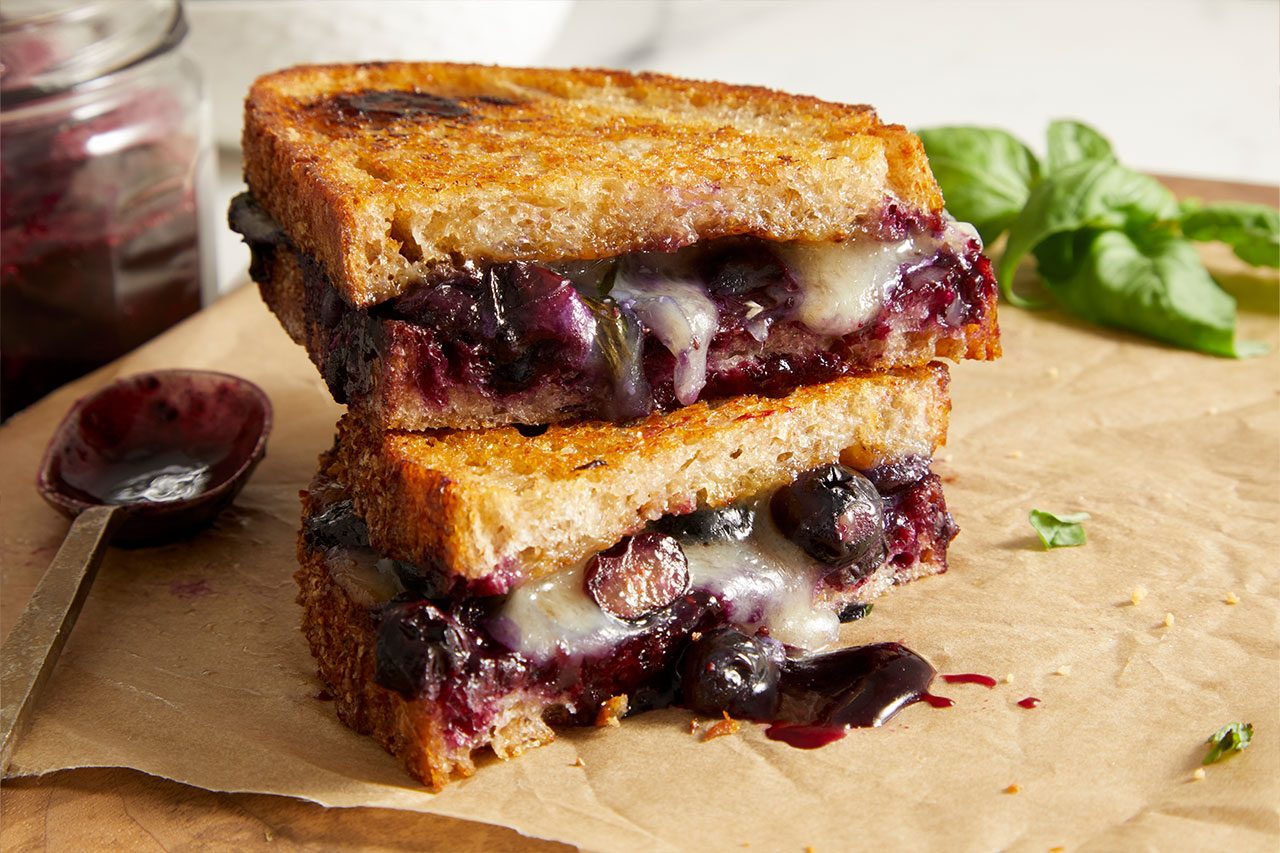 Close-up shot of Balsamic Blueberry Brie Grilled Cheese sandwiches stacked on parchment paper, featuring golden, crispy toasted bread layered with melted Brie cheese and warm blueberry compote, with fresh basil, a spoonful of blueberry compote, and a jar of compote softly blurred in the background.