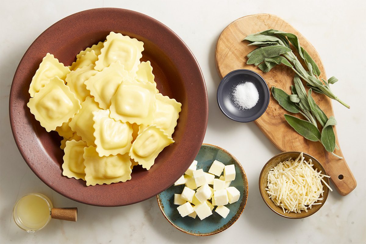 A bowl of uncooked ravioli sits beside sage leaves on a wooden board, a small dish of salt, a bowl of shredded cheese, cubed butter, and a cup of pale liquid on a white countertop.