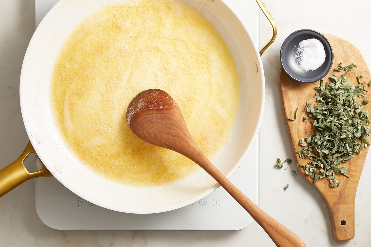 A wooden spoon rests in a white skillet with melted butter. On the side, a wooden cutting board holds chopped fresh herbs and a small dish of salt.
