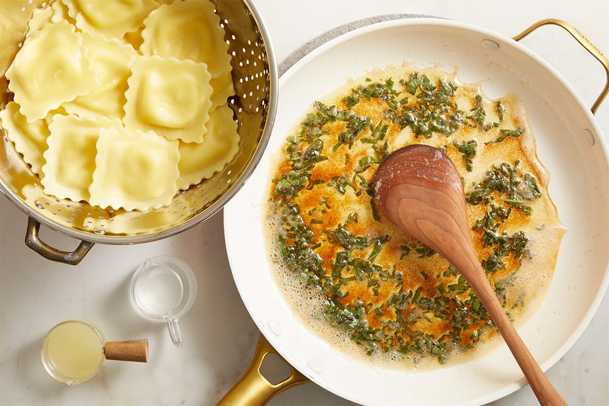 A skillet with browned butter and chopped herbs being stirred with a wooden spoon sits next to a colander of cooked ravioli and two small containers, one with grated cheese and one with liquid.