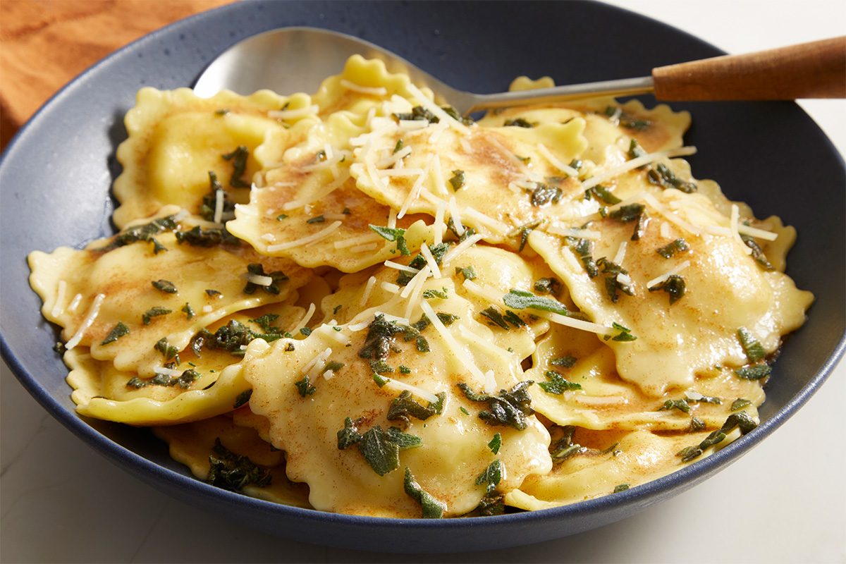 A bowl of ravioli topped with brown butter sauce, shredded Parmesan cheese, and fresh herbs, served with a spoon.