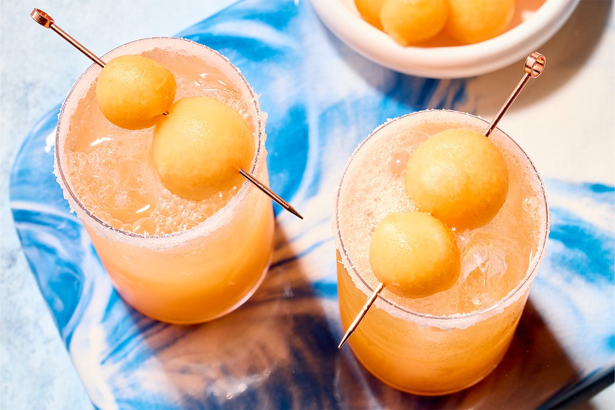 Overhead horizontal image of two vibrant yellow-orange cocktails, each garnished with melon balls on a metal skewer. Both glasses feature a sugared rim and are filled with a bubbly beverage and ice, resting on a blue and white patterned surface.