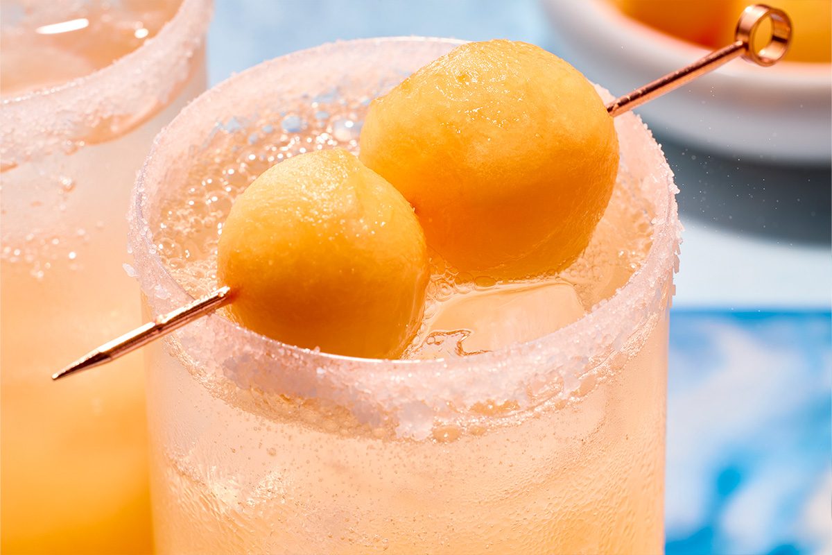 Eye Level horizontal image of a pale orange, bubbly drink in a glass with a sugar-coated rim. Two bright orange melon balls on a decorative skewer garnish the beverage, which also contains ice cubes.