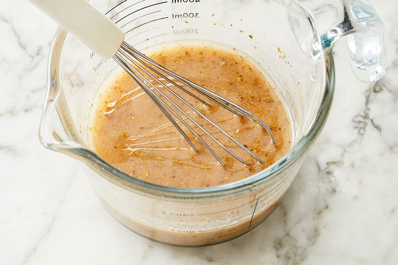 Overhead horizontal image of a whisk actively mixing ingredients in a glass measuring cup on a white marble surface. The liquid mixture, which appears to be a dressing or marinade, features visible layers of oil and specks of herbs and spices. The whisk is partially submerged in the liquid, combining the components.