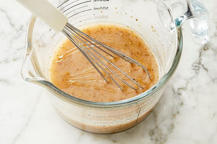 Overhead horizontal image of a whisk actively mixing ingredients in a glass measuring cup on a white marble surface. The liquid mixture, which appears to be a dressing or marinade, features visible layers of oil and specks of herbs and spices. The whisk is partially submerged in the liquid, combining the components.