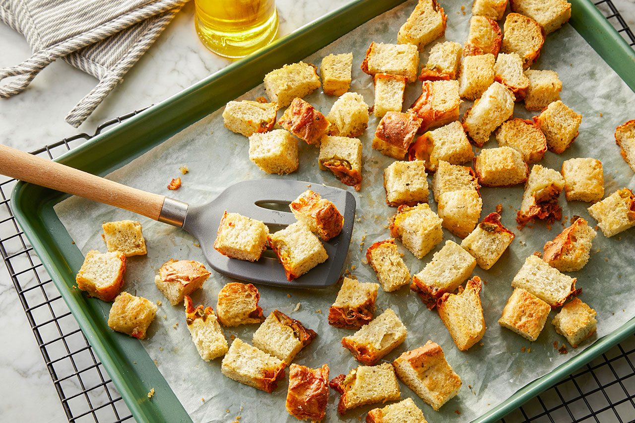 Overhead horizontal image of cubed bread pieces being toasted on a parchment-lined baking sheet. A gray spatula with a wooden handle rests among the golden-brown croutons. A glass of oil and a striped oven mitt are visible in the background.