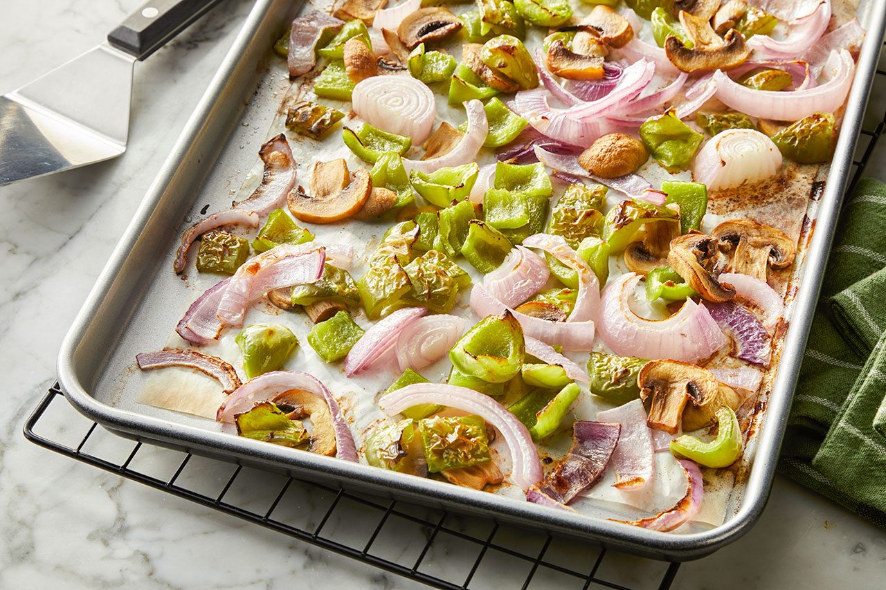 Overhead horizontal image of roasted green bell peppers, mushrooms, and onions on a parchment-lined baking sheet. The vegetables appear tender and slightly caramelized, ready for the next step in the recipe.