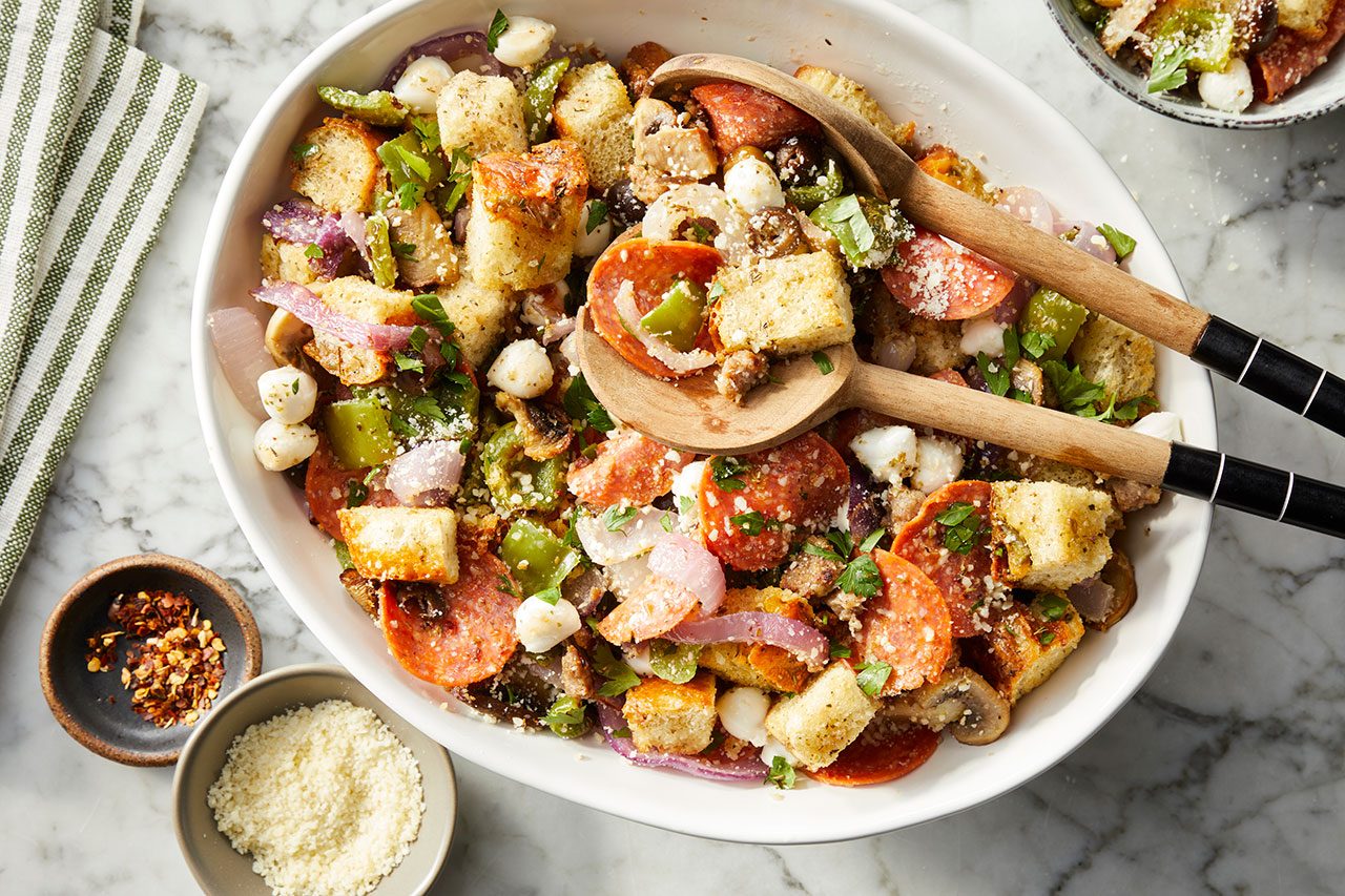 Overhead horizontal image of a large white oval serving bowl filled with a hearty salad featuring pepperoni, croutons, red onion, green bell pepper, mini mozzarella balls, and mushrooms, garnished with fresh herbs and grated Parmesan. Two wooden serving spoons are nestled within the dish, with small bowls of chili flakes and grated Parmesan alongside.