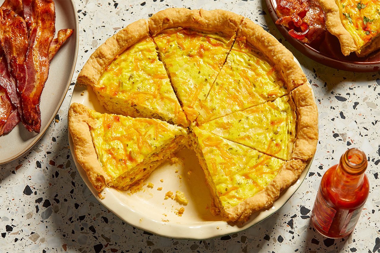 Overhead horizontal image of a freshly baked Chile Relleno Quiche, cut into wedges, presented in a pie dish on a speckled countertop. One slice of quiche has been removed, revealing the cheesy, herb-flecked filling. A bottle of hot sauce and a plate of crispy bacon are visible on the sides.