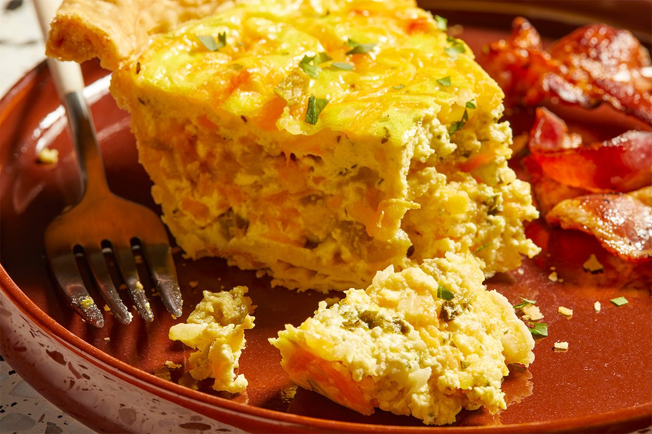 Eye Level horizontal image of a slice of Chile Relleno Quiche served on a reddish-brown plate. A piece of the quiche has flaked off onto the plate next to a fork, revealing its savory filling with green chiles and cheese. Crispy bacon pieces are visible on the right side of the plate.