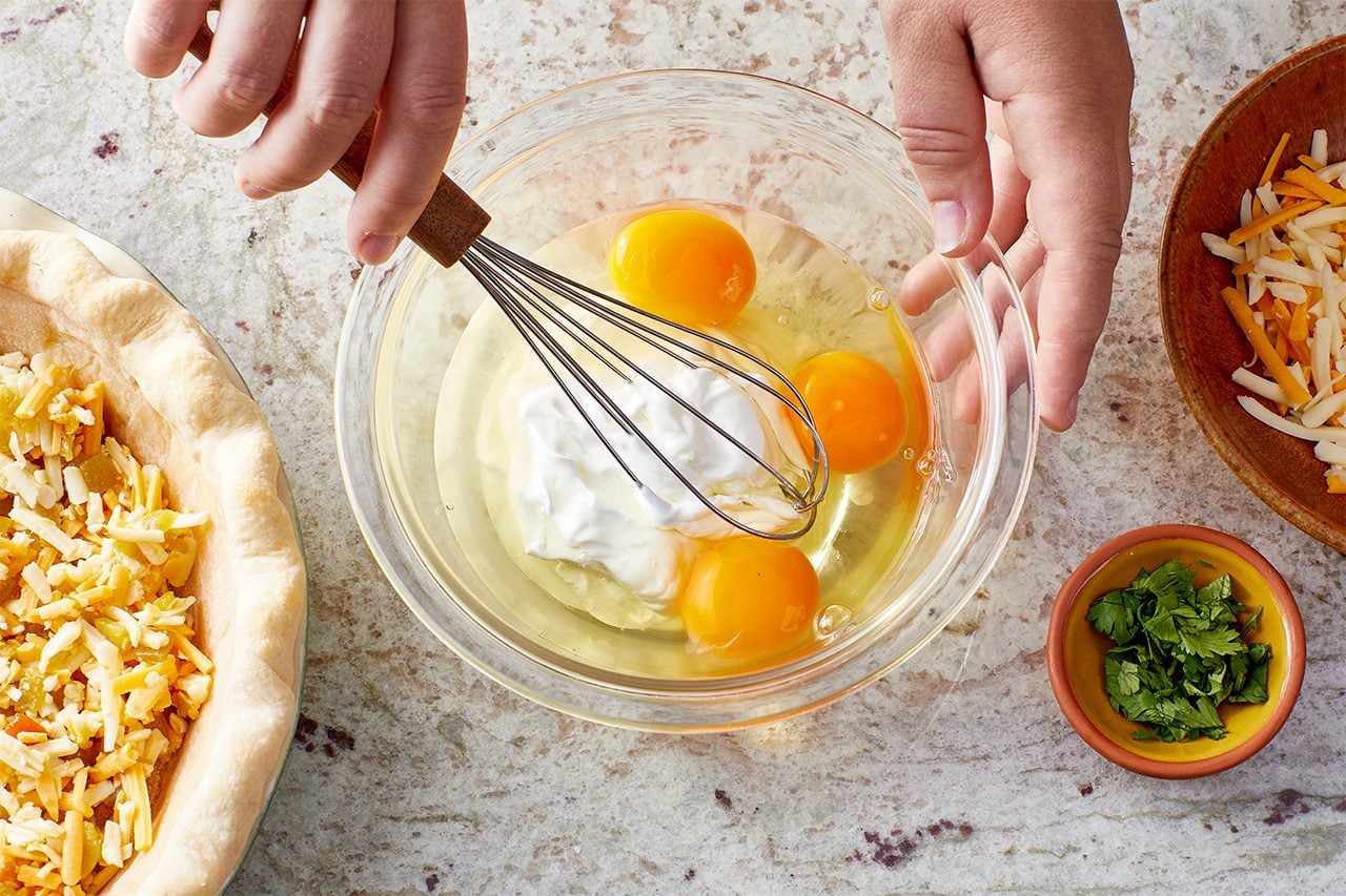 Overhead horizontal image of hands holding a whisk in a clear bowl containing cracked eggs and sour cream. To the left, a pie crust is filled with shredded Monterey Jack and cheddar cheeses and chopped green chiles, with a small bowl of minced fresh cilantro to the right.
