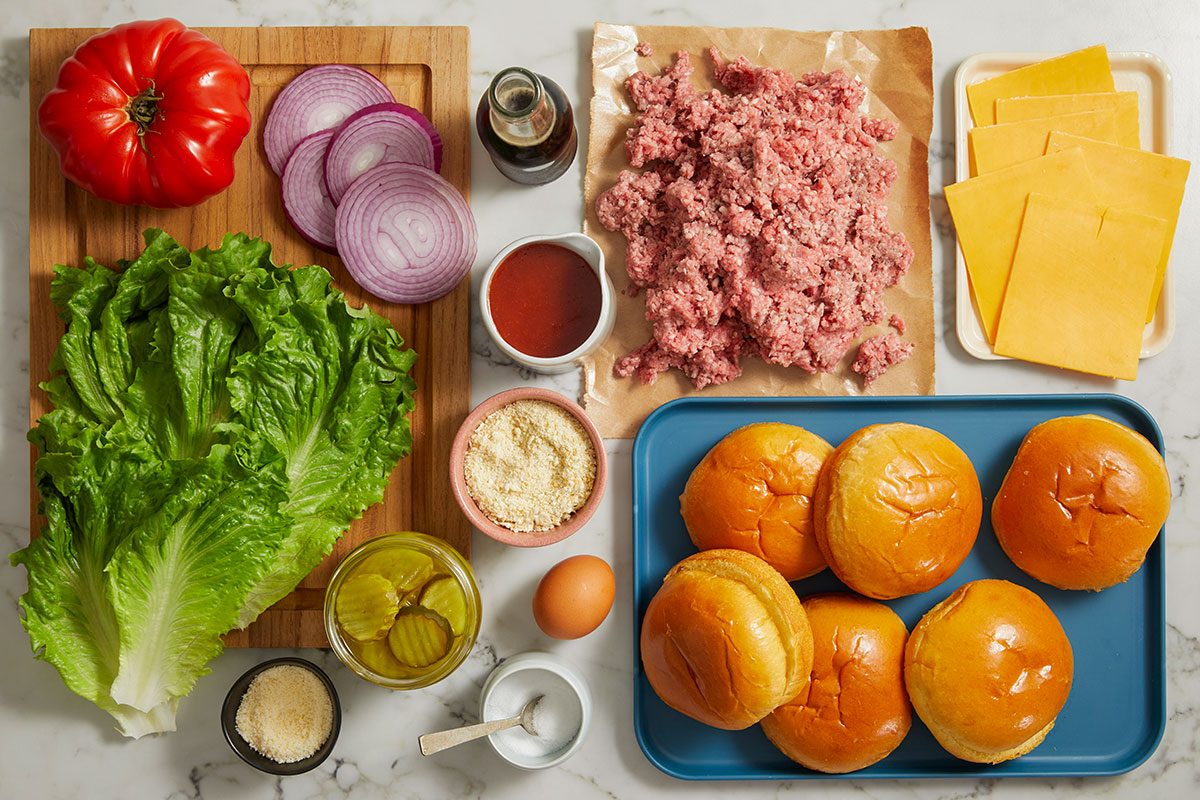 Overhead shot of fresh burger ingredients, including ground beef, buns, cheddar cheese, lettuce, tomato, red onion, pickles, ketchup, seasonings, an egg, and a bottle of cola, neatly arranged on wooden boards atop a light marble countertop; ready for preparing Cola Burgers;