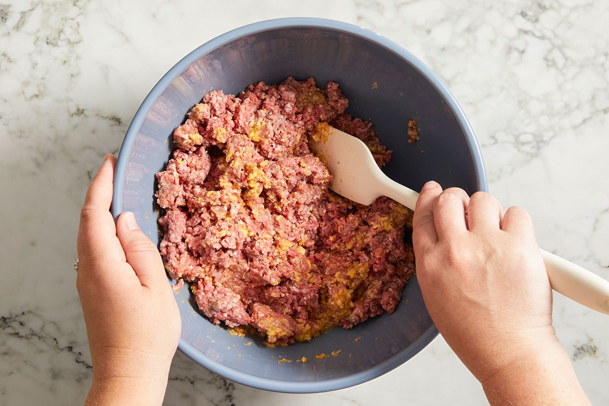 Overhead shot of hands mixing seasoned ground beef in a large bowl with a white spatula on a light marble countertop;