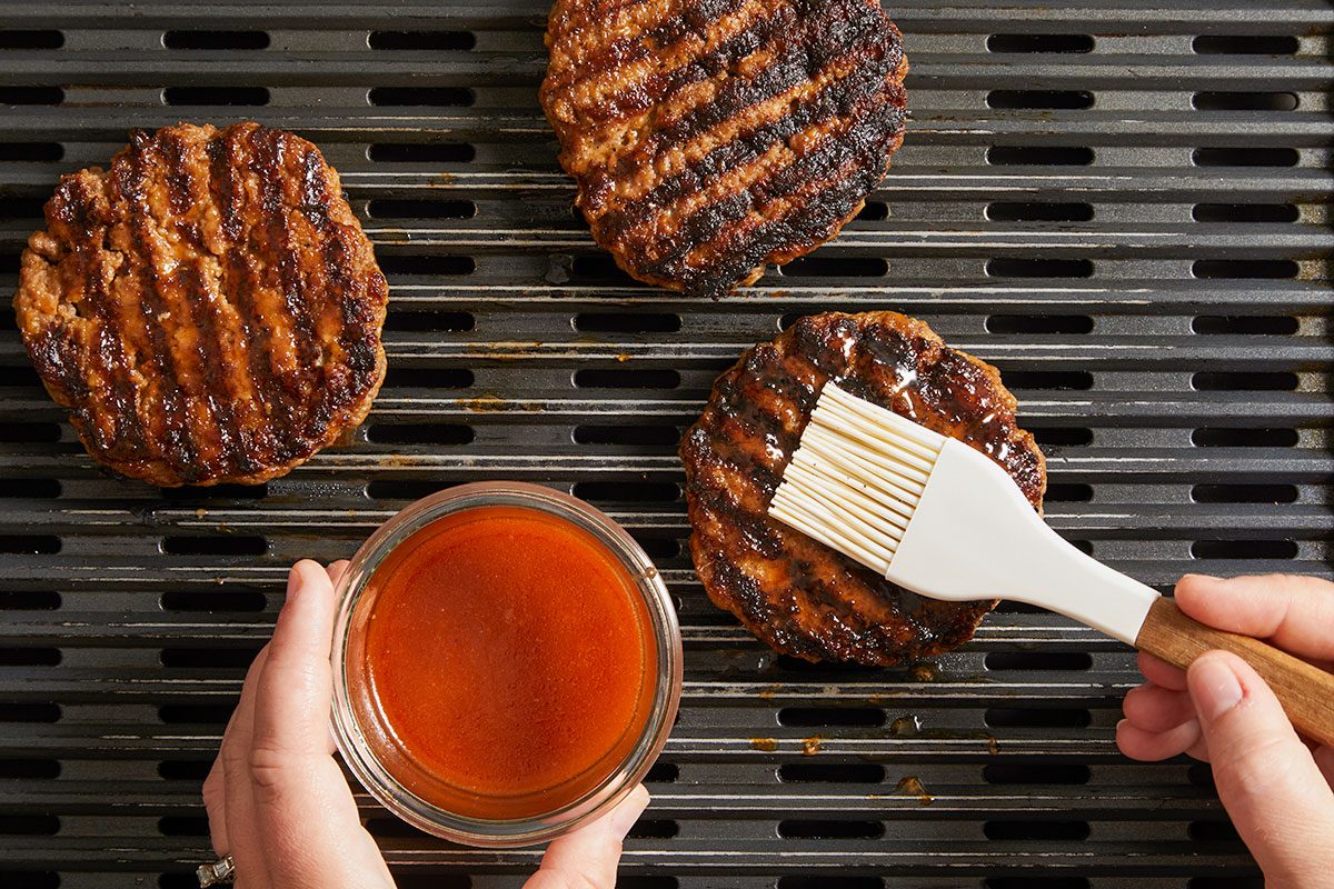 Overhead shot of grilled burger patties on a grill being brushed with cola glaze, with one hand holding a bowl of sauce;