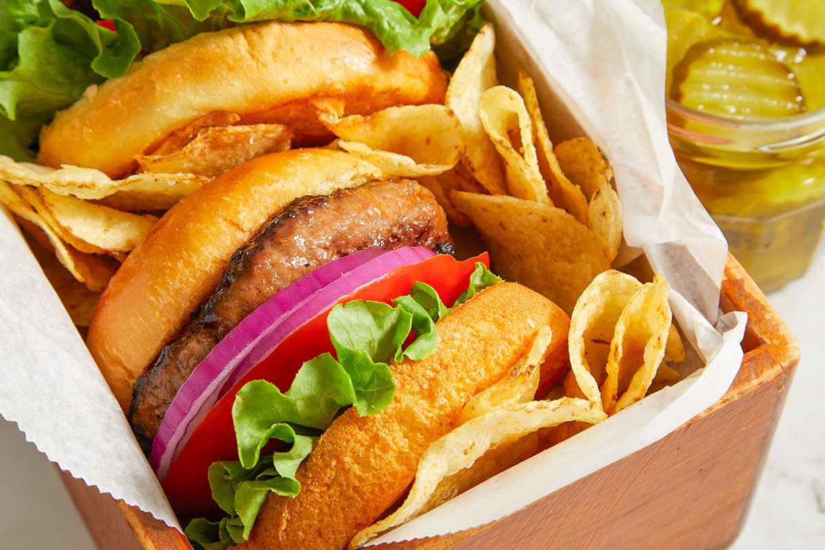 Closeup shot of Cola Burgers layered with grilled beef patties, crisp lettuce, red onion slices, and soft toasted buns, served with crispy potato chips in a parchment-lined wooden tray on a light marble countertop;