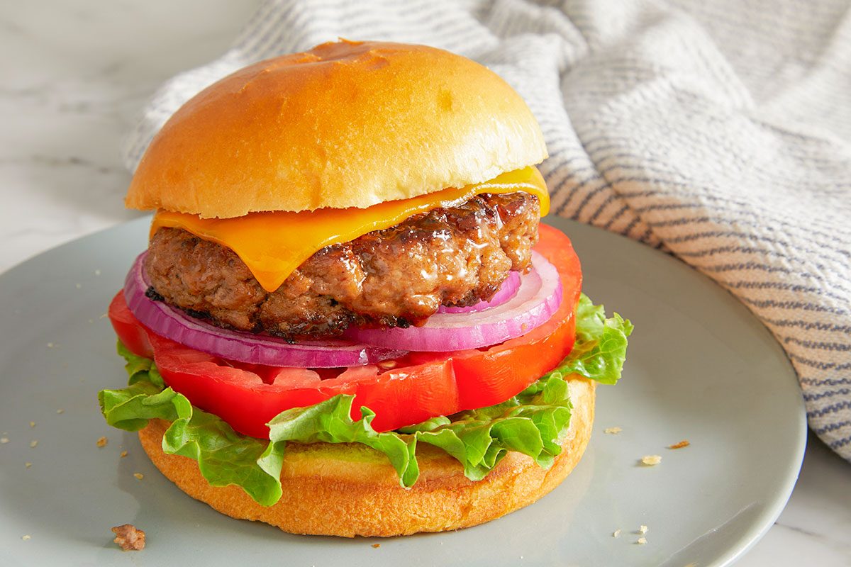 Close-up shot of a finished Cola Burger layered with a grilled beef patty, melted cheddar cheese, fresh tomato, red onion, and crisp lettuce on a soft toasted bun, served on a grey plate with a light marble countertop and a neutral textured cloth in the background;