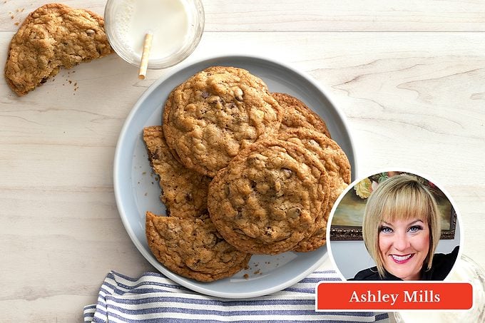 A plate of oatmeal cookies sits on a white table with a glass of milk and a striped napkin. An inset features a smiling woman labeled “Ashley Mills.”.