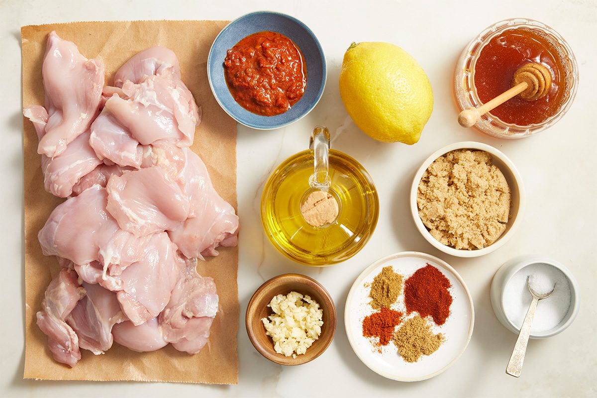 Overhead shot of raw chicken pieces arranged on parchment paper alongside bowls of olive oil, honey, paprika, garlic, spices, brown sugar, lemon, tomato paste, and salt, neatly organized on a light surface