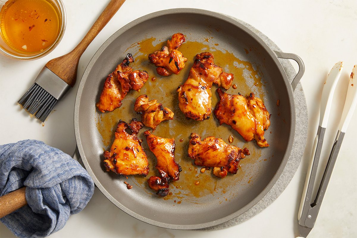 Overhead shot of caramelized chicken pieces roasting in a skillet, coated in a glossy honey-spice glaze with lightly charred edge;. A bowl of extra glaze, a pastry brush, kitchen towel, and metal tongs are arranged nearby on a light surface;