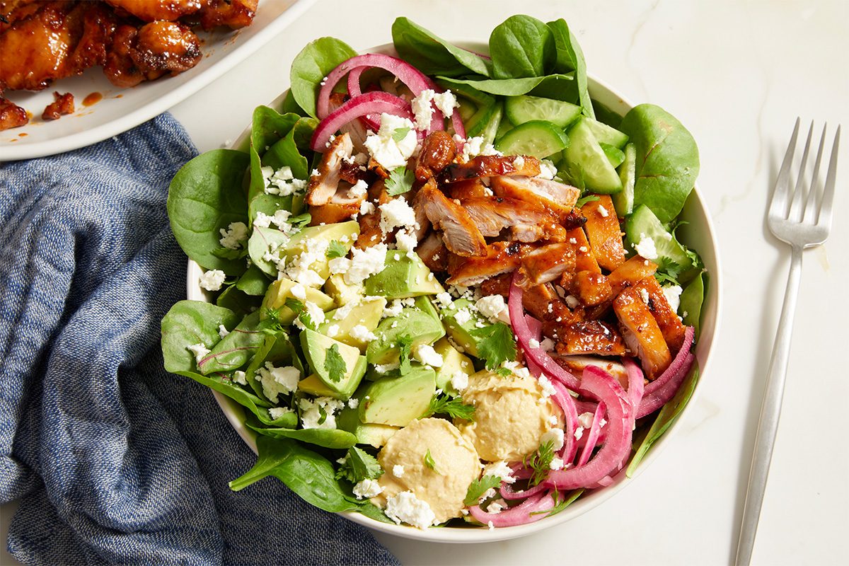 Top view shot of Copycat Cava Harissa Honey Chicken served over a fresh salad of spinach, cucumbers, avocado, hummus, pickled red onions, feta cheese, and herbs in a white bowl, with a fork and roasted chicken platter nearby;